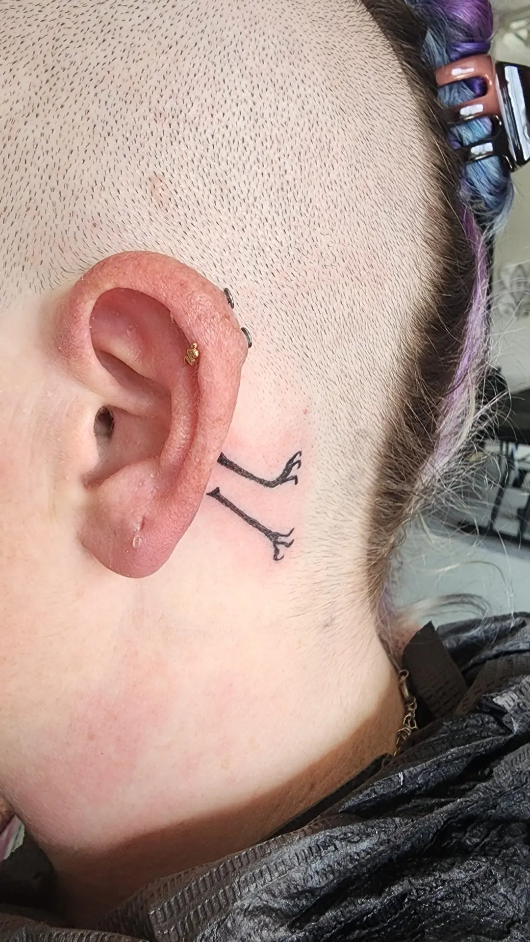 Two tiny black bird legs tattooed behind an ear, as if a bird is hiding behind it