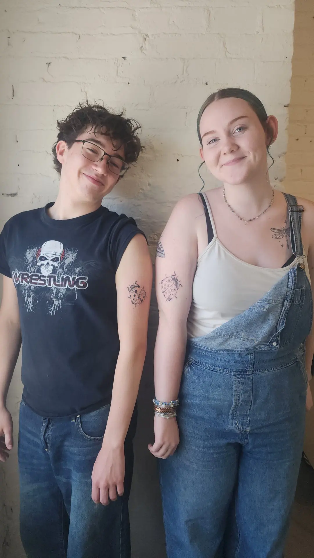 Two friends smiling against a white brick wall, each showing a fresh ladybug tattoo on the arm