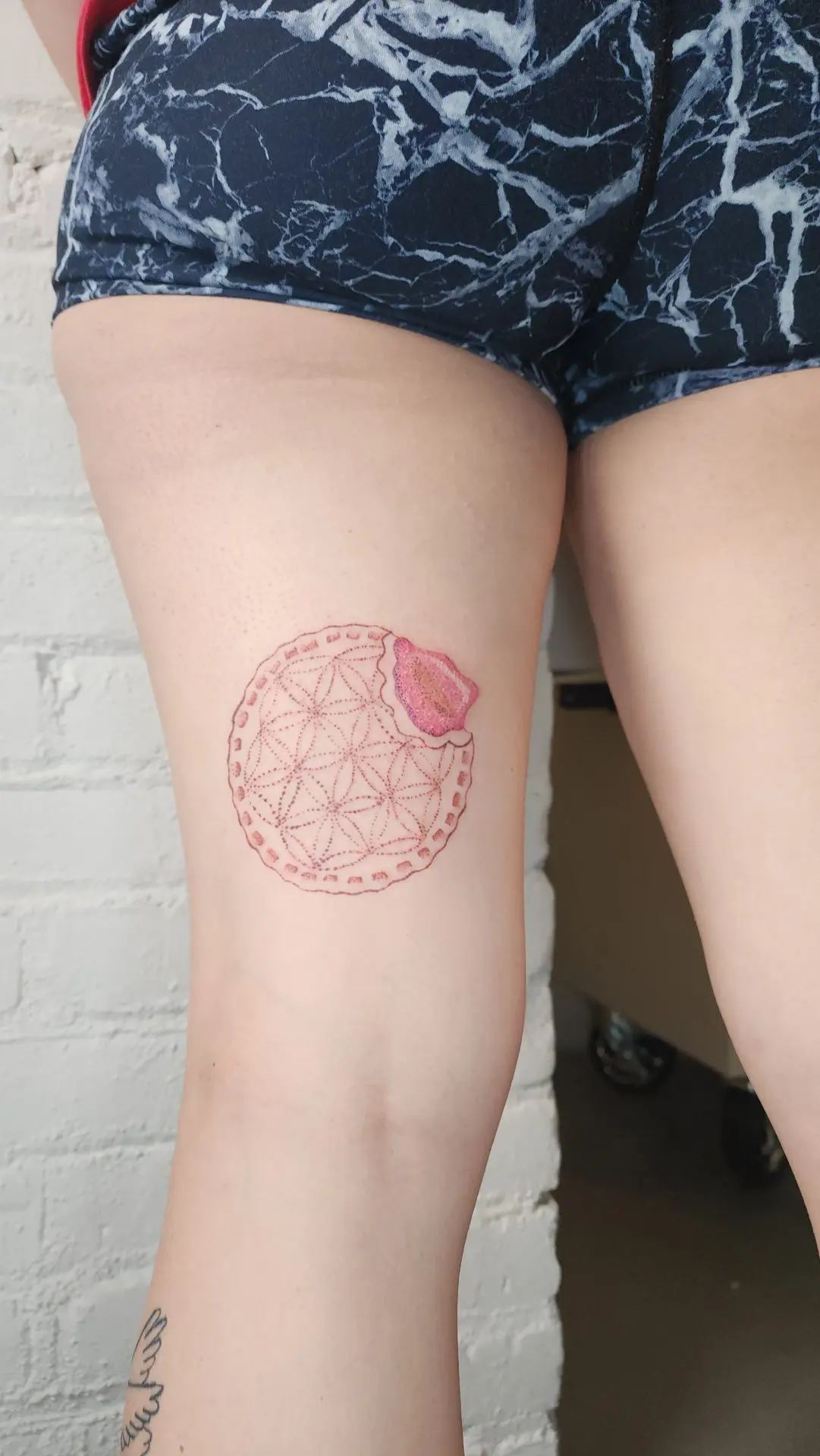 Flower-of-life pattern in dotwork shaped like a bitten cookie, with a pink color-packed bite mark, on the back of a thigh